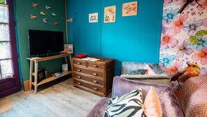 A room with a television on a wooden stand, a dresser, a bed with patterned bedding, and wall art including a bird and floral designs.