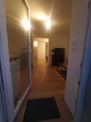 Un couloir mène à une pièce dotée d’une porte-fenêtre coulissante, d’un petit tapis, d’un téléviseur et d’un meuble.