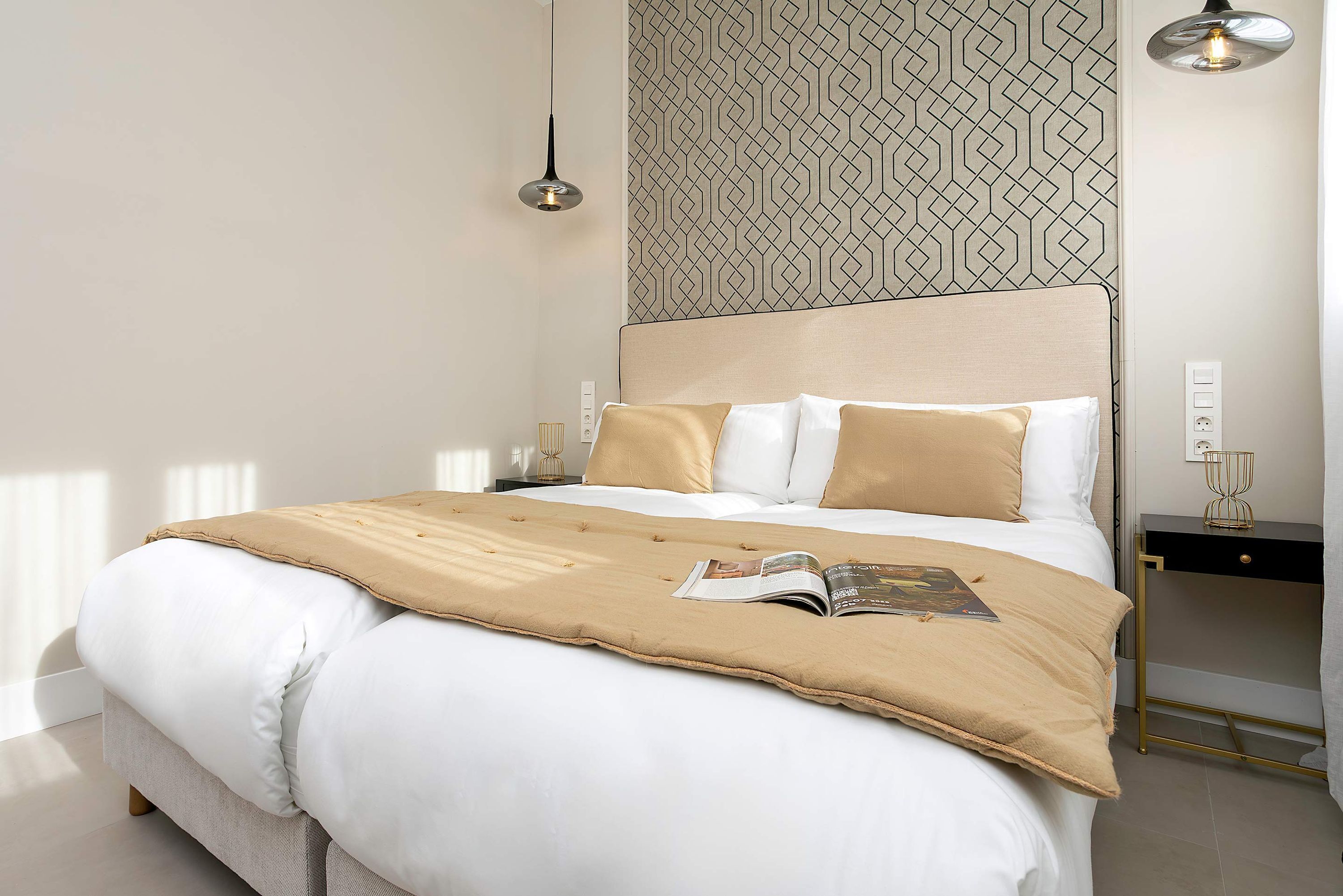 A neatly made bed with a beige throw blanket and pillows, a bedside table with a lamp, and a patterned wall.