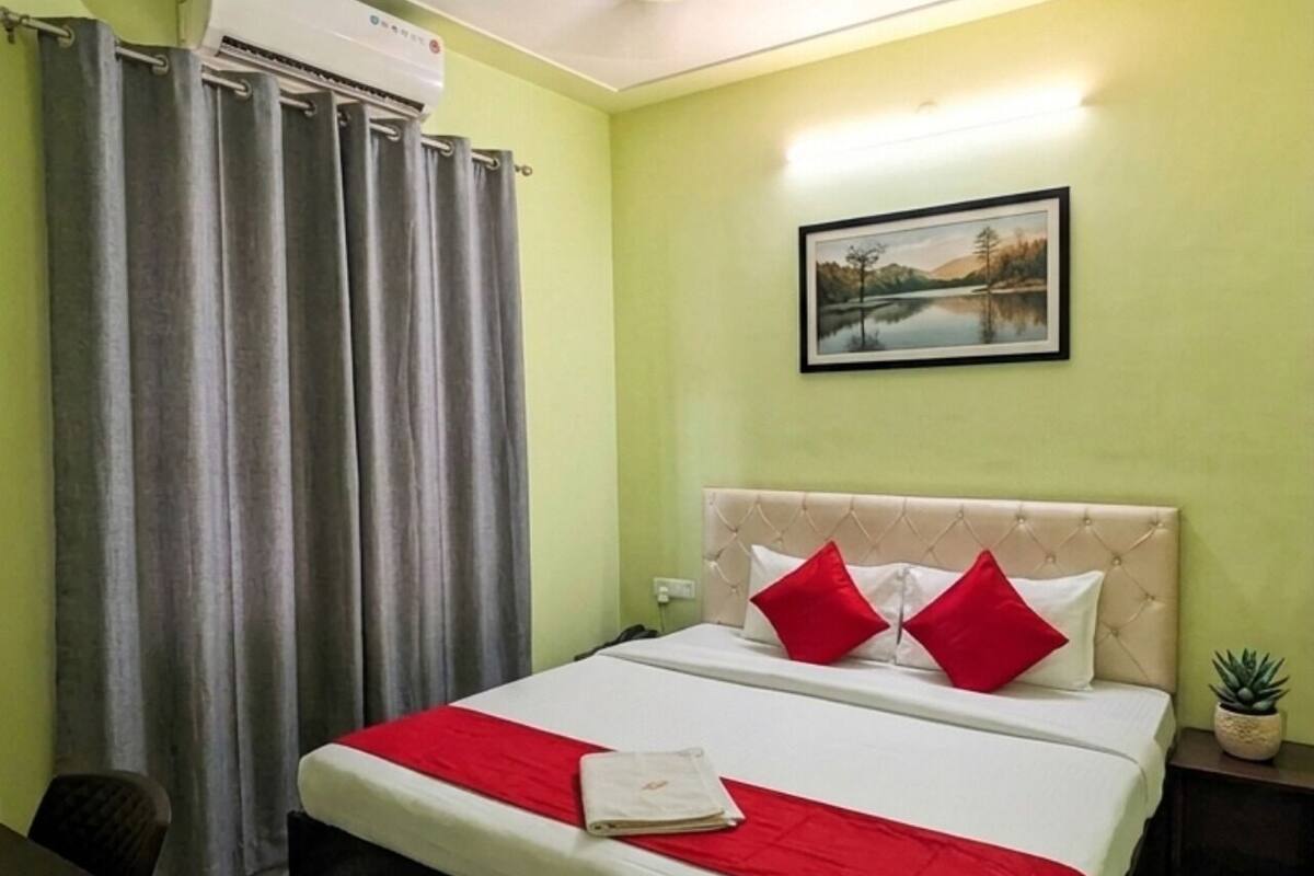 A hotel room with a bed, grey curtains, a wall-mounted air conditioner, a framed picture, and a small potted plant.