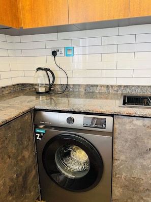 Eine kompakte Waschmaschine und ein Trockner mit Bedienfeld, ein Wasserkocher auf der Arbeitsplatte und eine geflieste Spritzschutzwand.