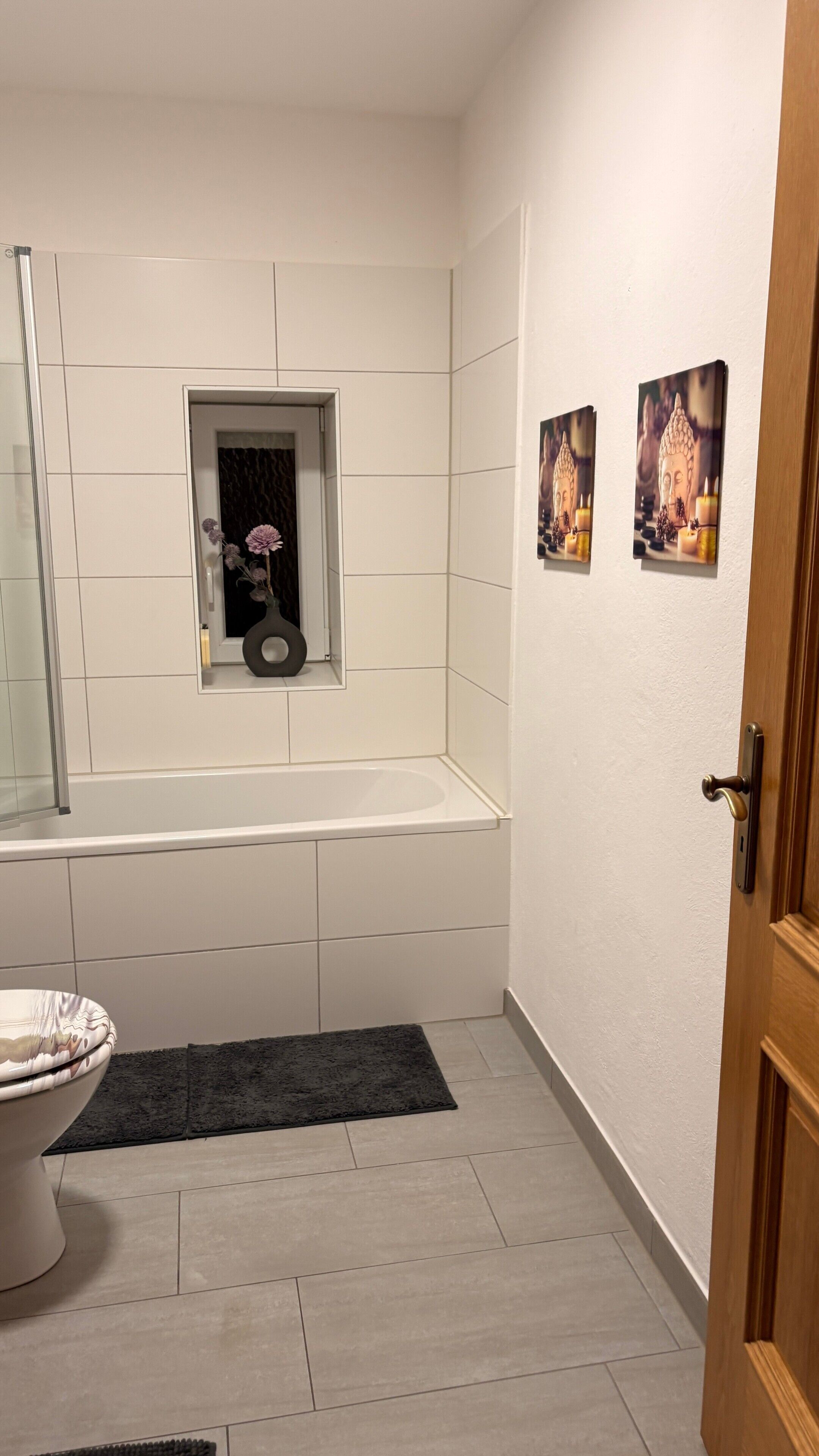 Une salle de bain avec une baignoire, des toilettes, une petite fenêtre décorée de fleurs et deux tableaux accrochés au mur.