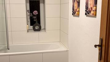 Une salle de bain avec une baignoire, des toilettes, une petite fenêtre décorée de fleurs et deux tableaux accrochés au mur.