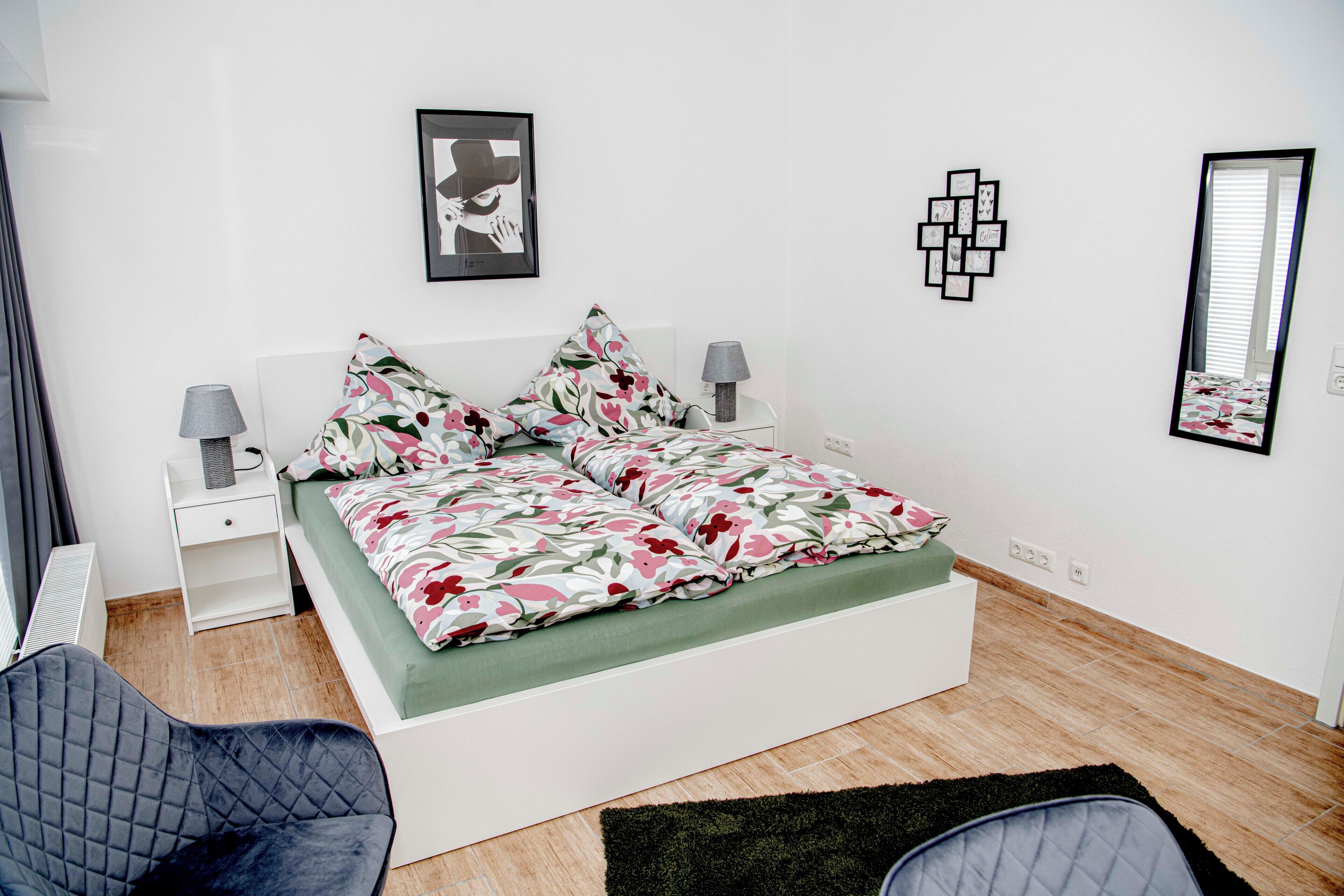 A bedroom with a bed, two bedside tables, a chair, a mirror, and wall decorations.