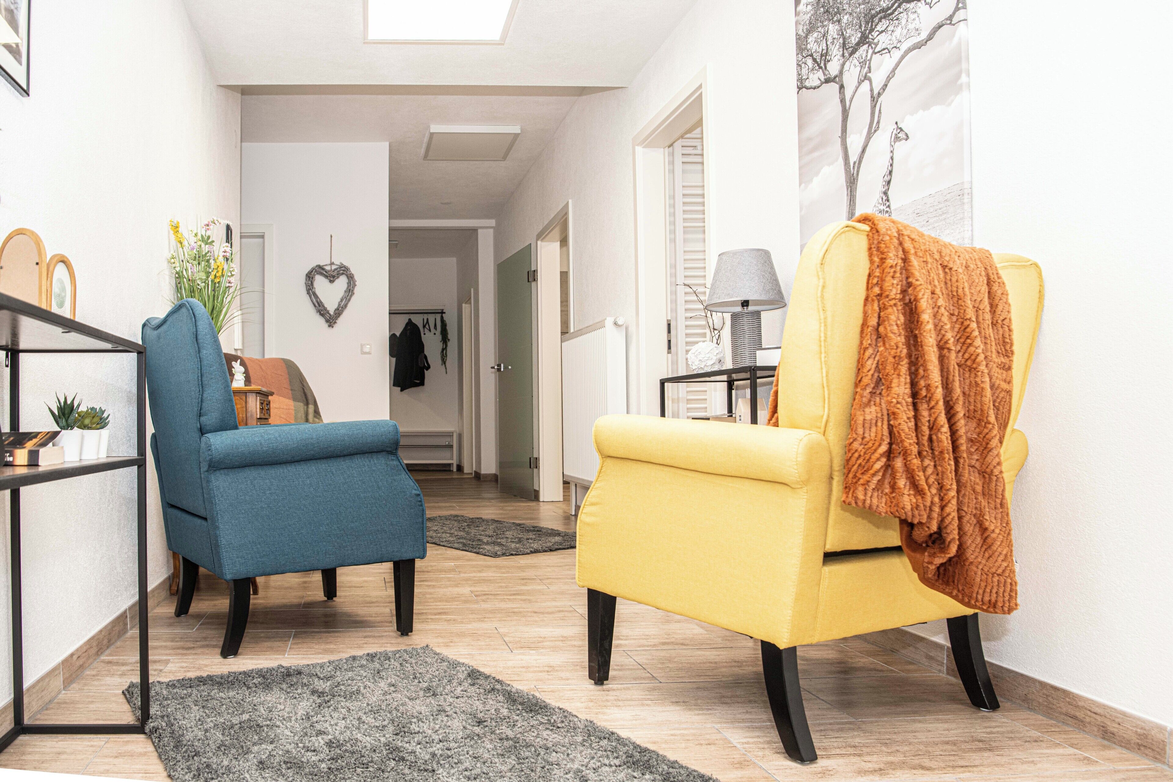 Un salon avec un fauteuil bleu, un fauteuil jaune, une petite table avec une lampe et un tableau accroché au mur.
