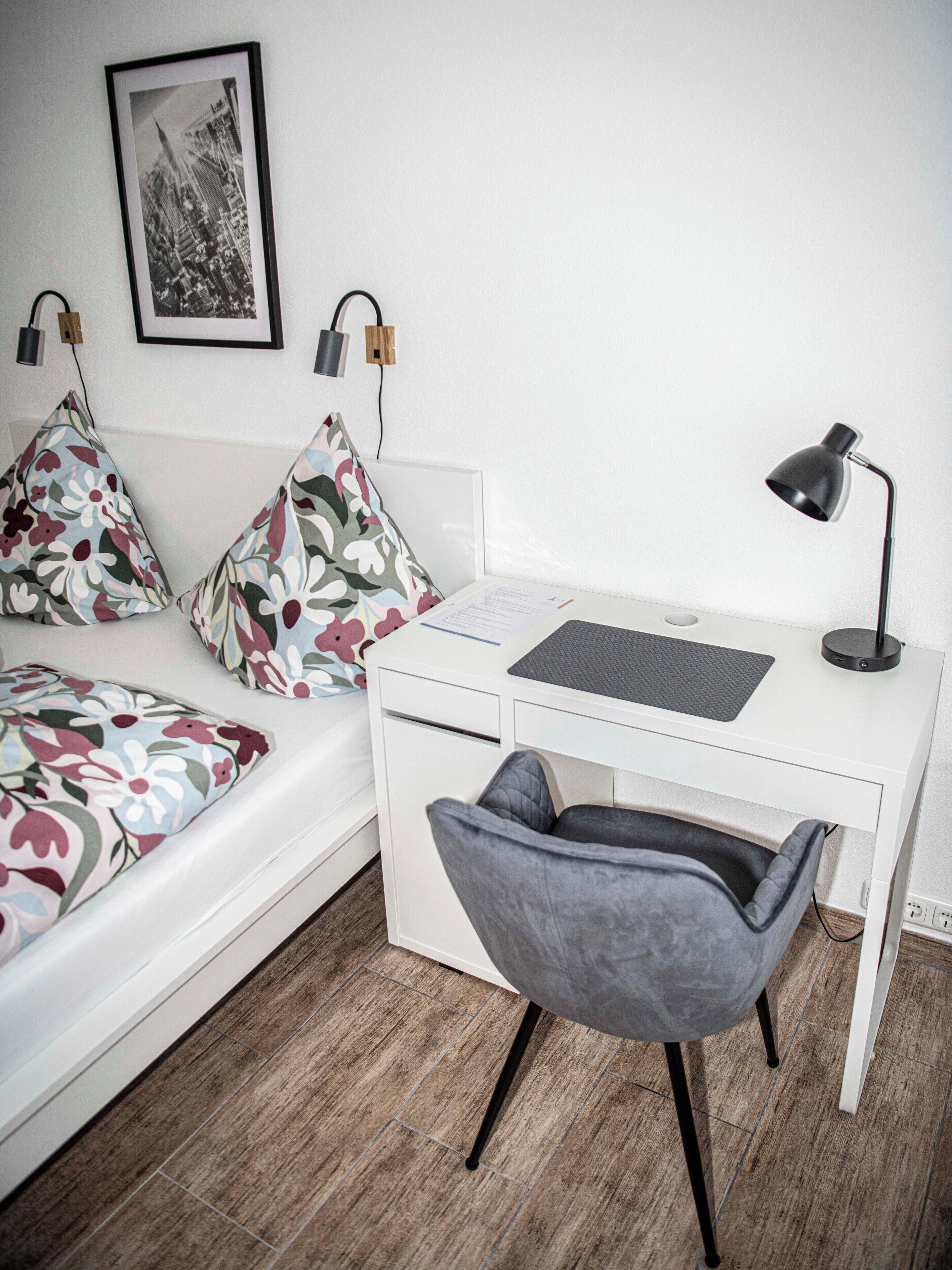 A neatly arranged bedroom with a bed, a desk, a chair, and a desk lamp.