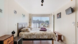 Un dormitorio con cama, escritorio, silla, ventana con vistas y televisor montado en la pared.