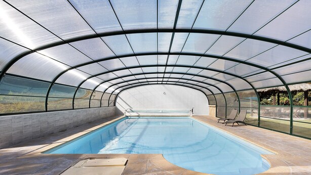 Una piscina interior con techo curvo, paredes de vidrio transparente y suelo de baldosas.