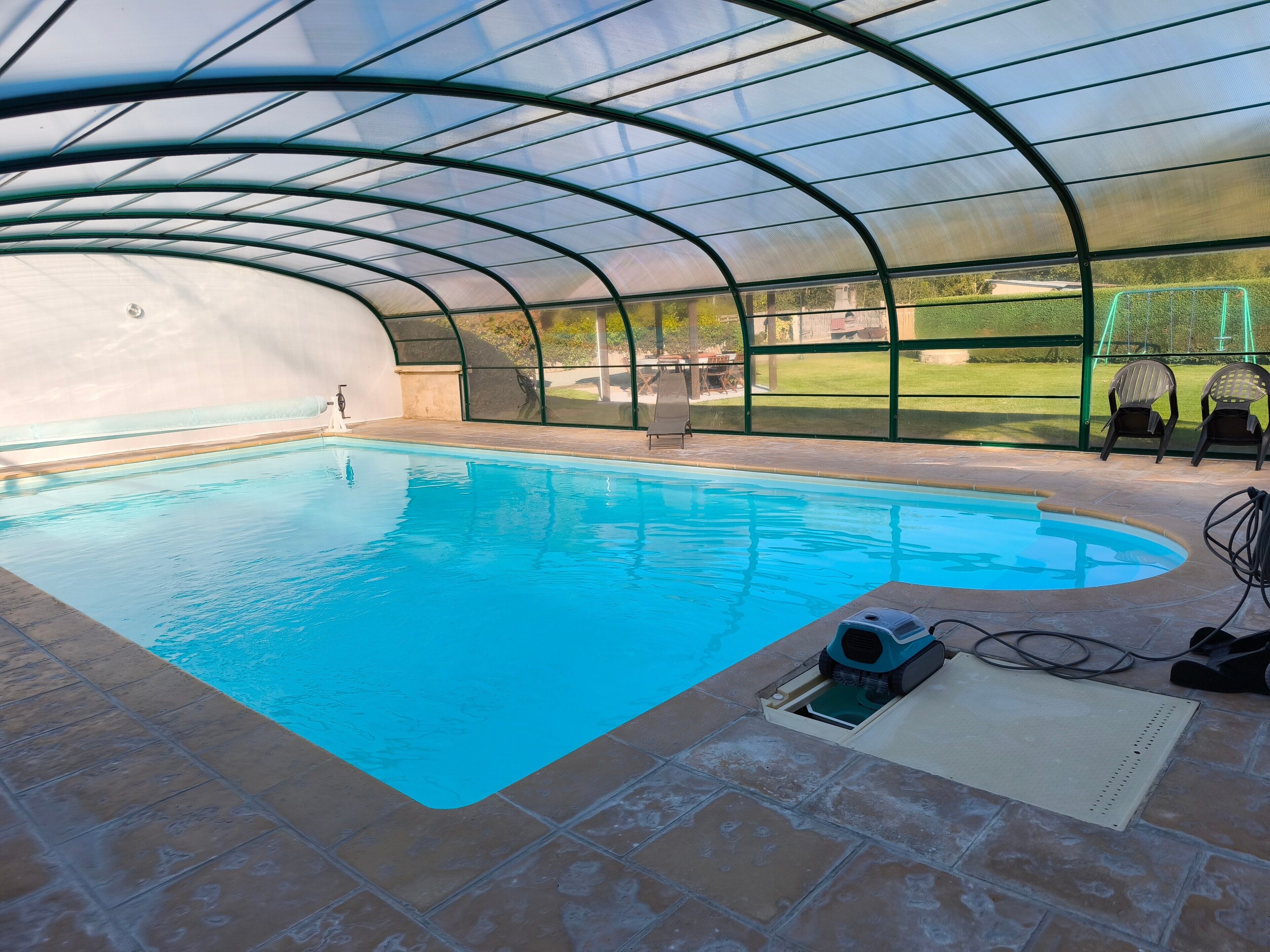 An indoor swimming pool with a curved roof and clear glass walls, surrounded by a patio area with chairs.