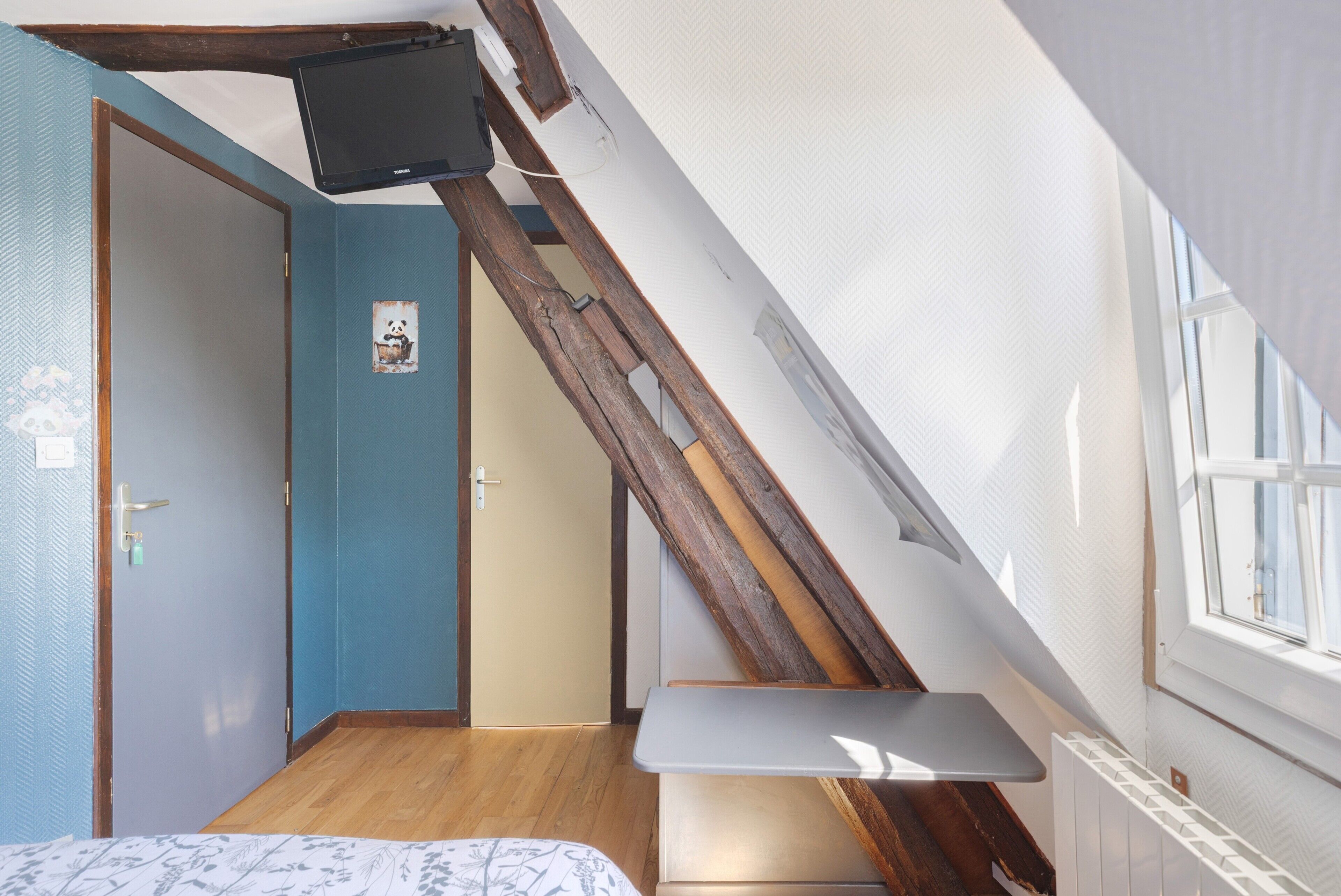 Une pièce avec un plafond en pente, un lit, une petite table, un téléviseur fixé au plafond et une fenêtre.