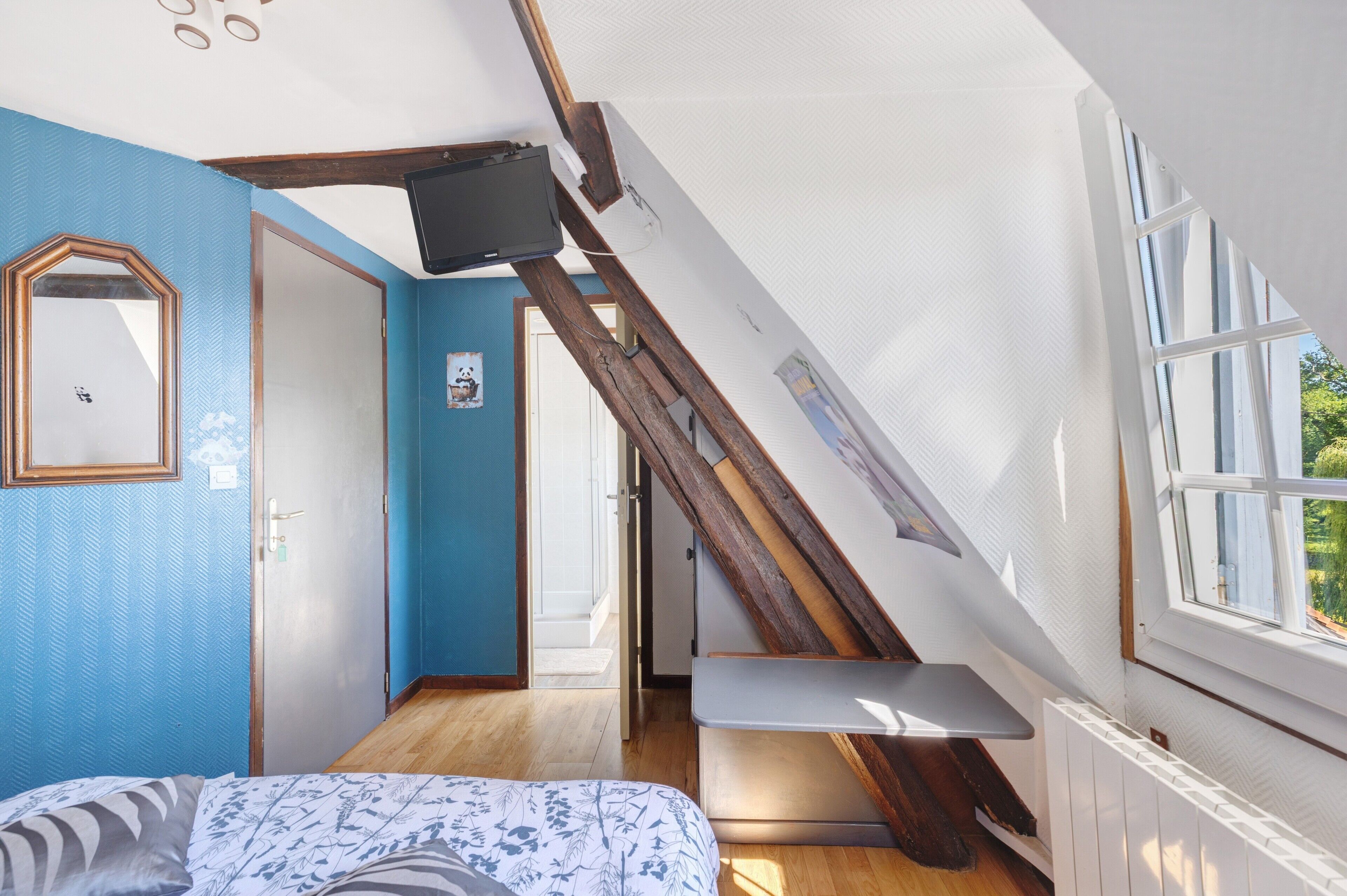 Quarto com teto inclinado, cama com roupa de cama estampada, escada de madeira, televisão e janela com vista para o verde.