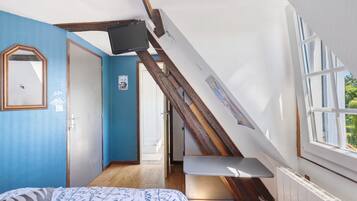 Quarto com teto inclinado, cama com roupa de cama estampada, escada de madeira, televisão e janela com vista para o verde.