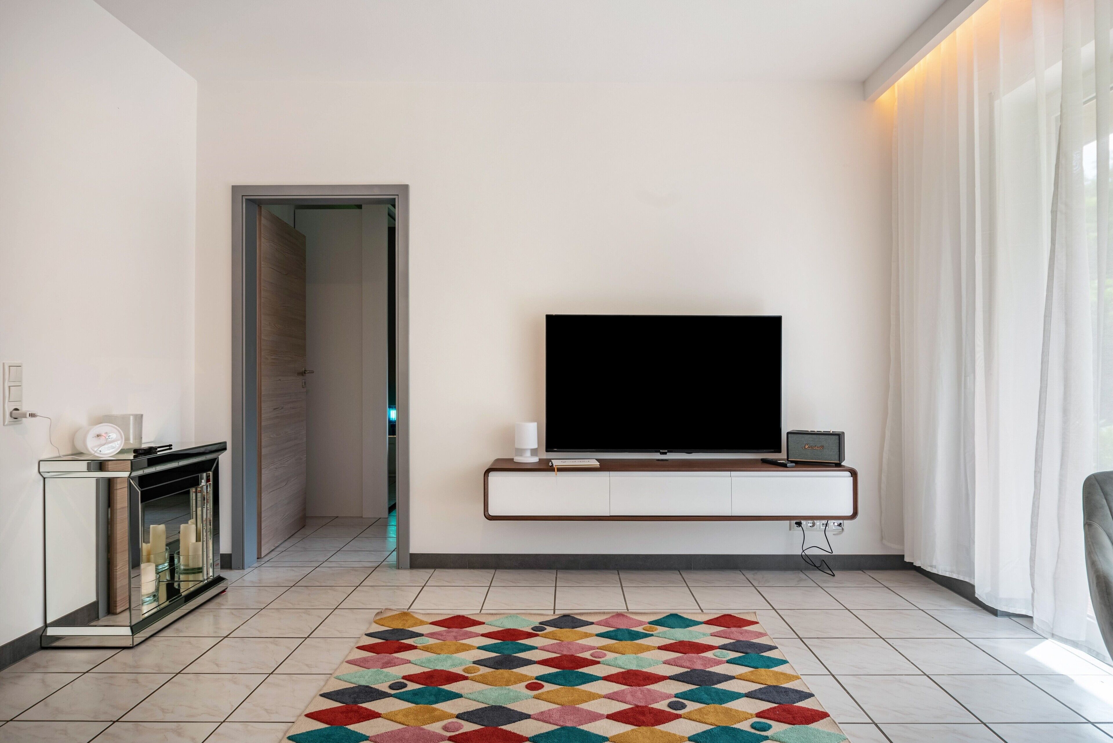A modern living room with a flat-screen TV mounted on a wall, a fireplace with a glass enclosure, and a colorful geometric rug.