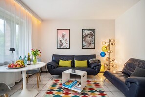 A modern living room with a black leather sofa, a white coffee table, and a colorful rug. There are framed artworks on the wall, a lamp, and a small table with fruits and drinks. - Apartment 'Ferienwohnung Für 2 Personen' with Wi-Fi (Traben-Trarbach)