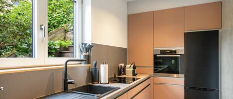 A modern kitchen with a built-in oven, a stainless steel refrigerator, and a window overlooking greenery.