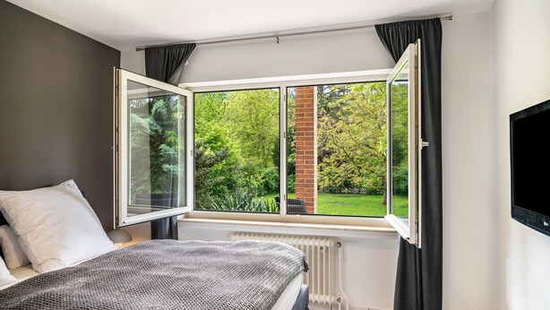 A bedroom with a large window, a radiator, a bed with a blanket, and a TV mounted on the wall.
