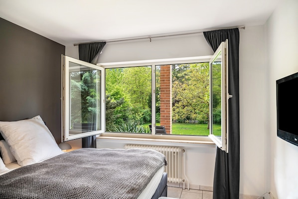 Un dormitorio con un ventanal grande, un radiador, una cama con una manta y un televisor montado en la pared.