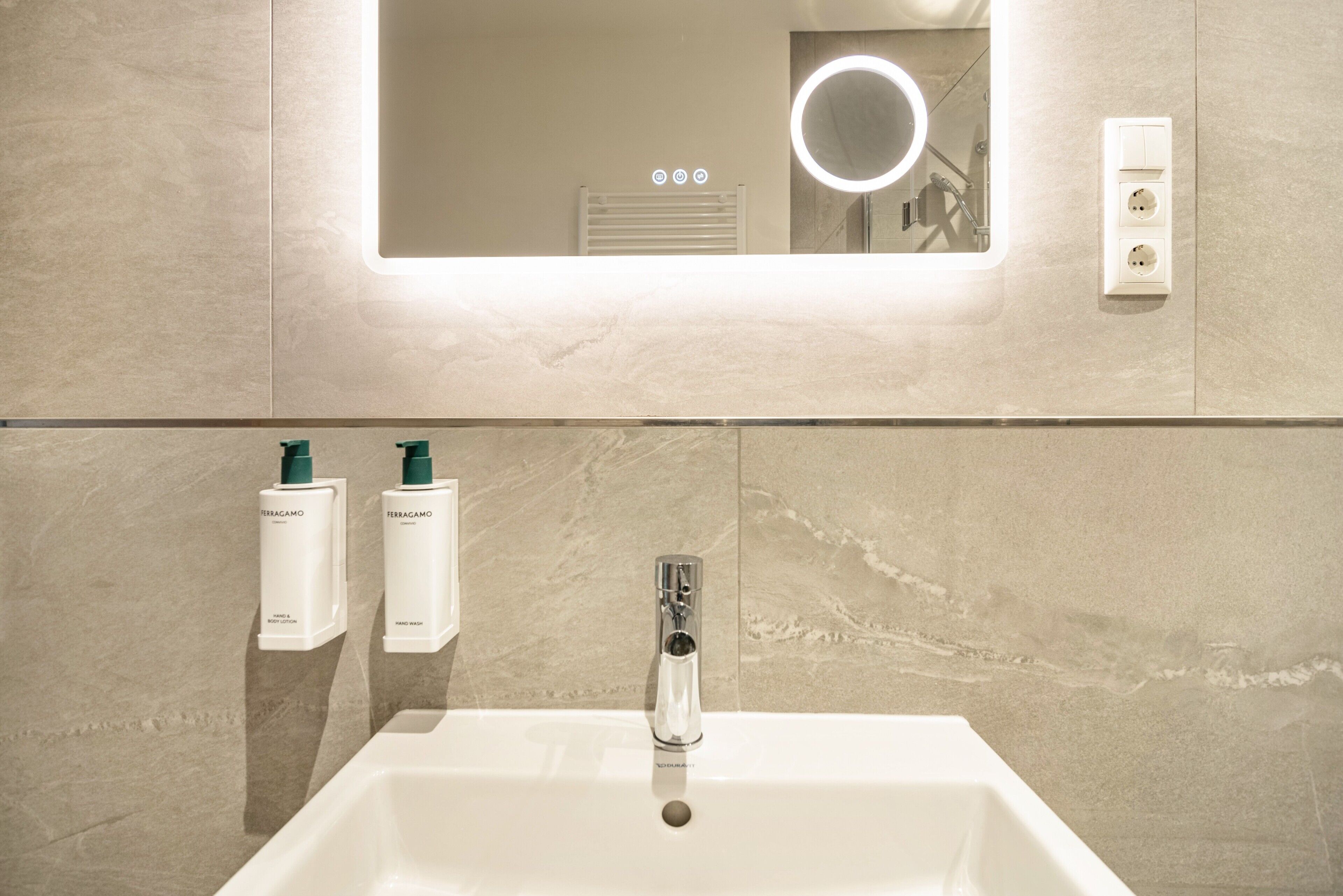 A bathroom with a white sink, a mirror with a circular light, and two bottles of Ferragamo hand lotion.