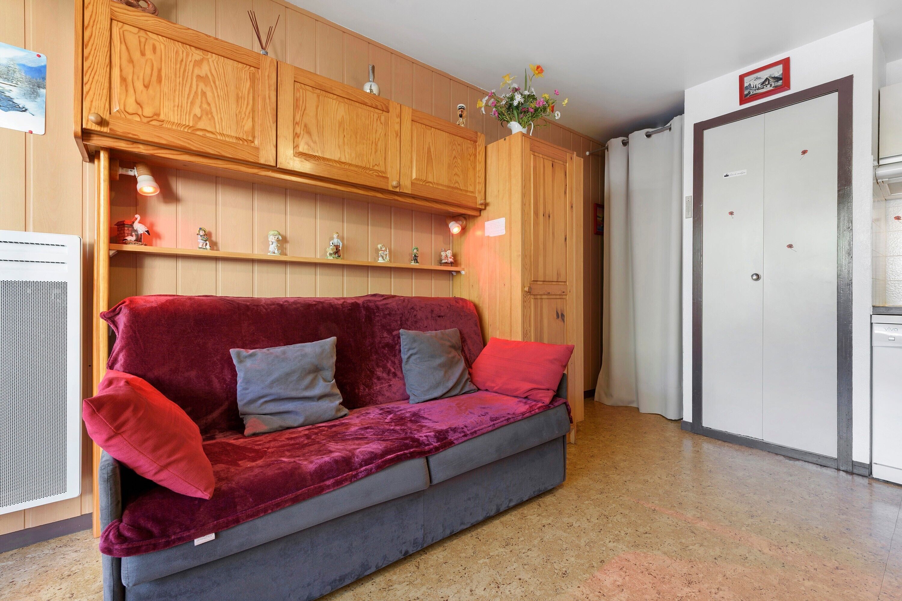 A room with a red velvet sofa, wooden cabinets, a white door, and a small shelf with decorative items.