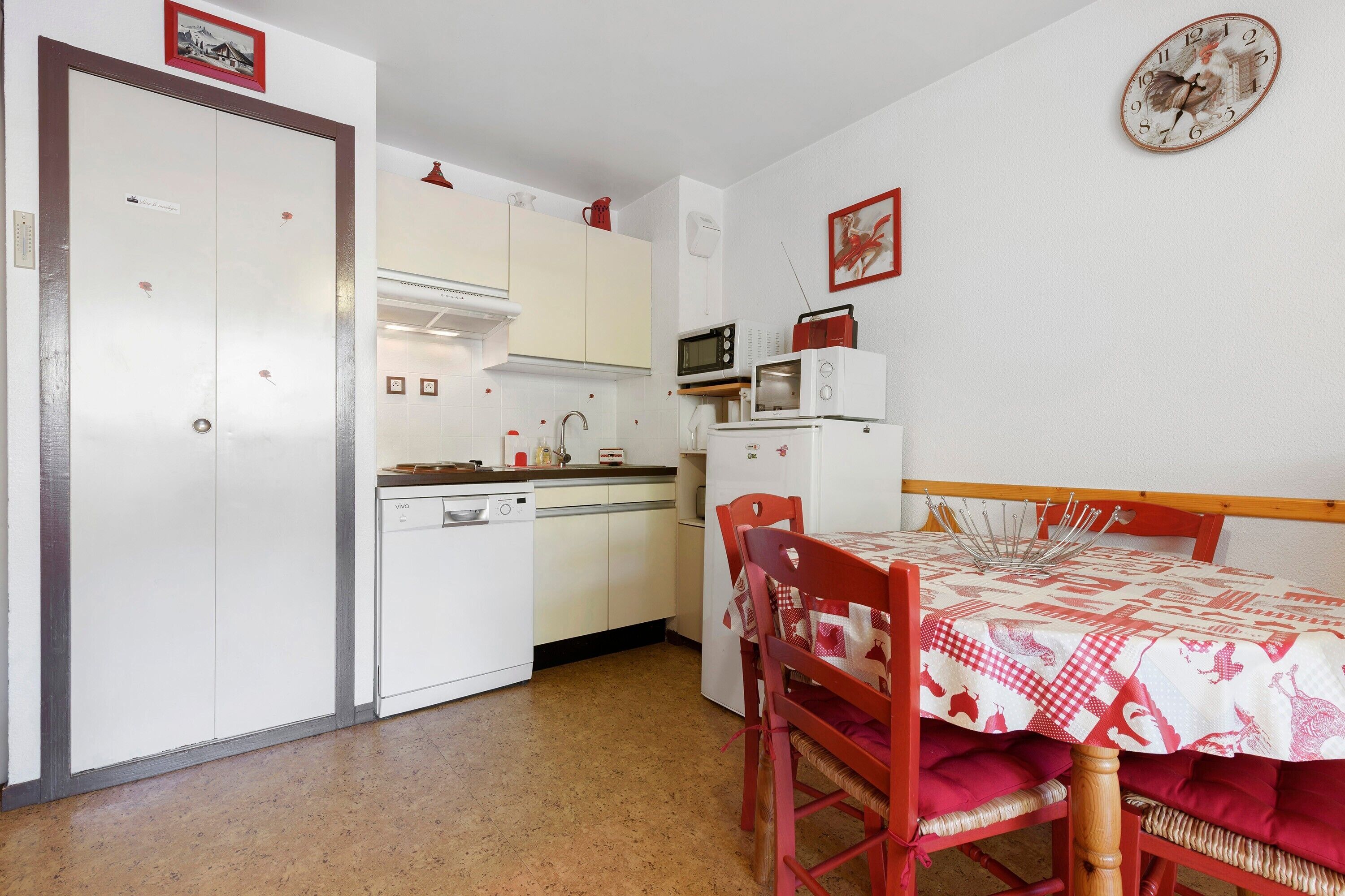 Een compacte keuken met witte kastjes, een magnetron, en een eethoek met een rode tafel en stoelen.