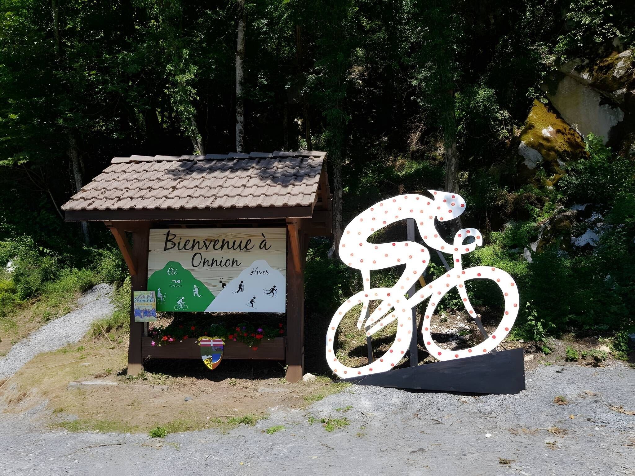 Un panneau d’accueil avec un symbole de vélo, un petit abri et un sentier menant à une zone boisée.