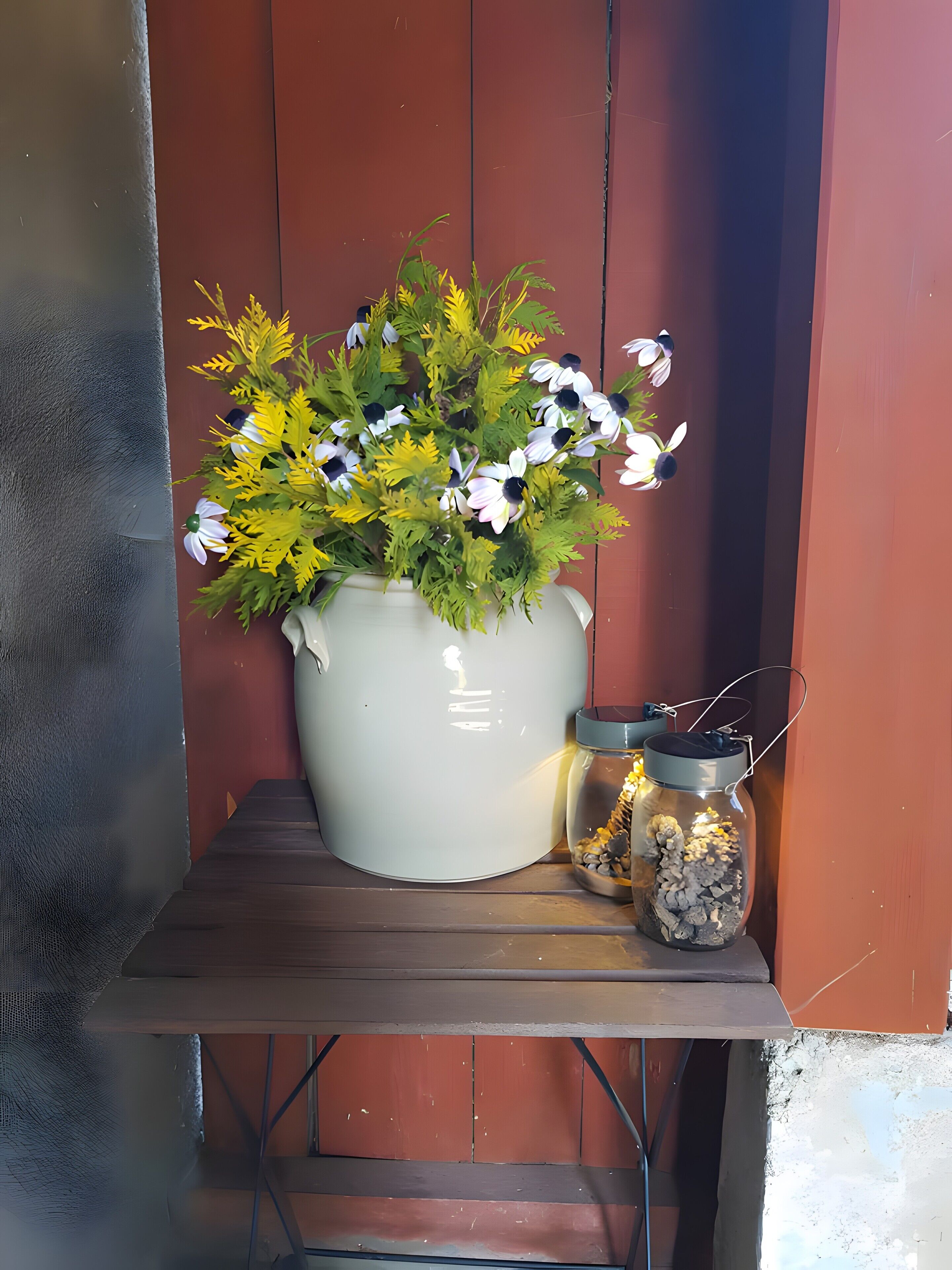 Un pot blanc orné de fleurs et un bocal en verre rempli de petits cailloux, posés sur une table en bois devant un mur rouge.
