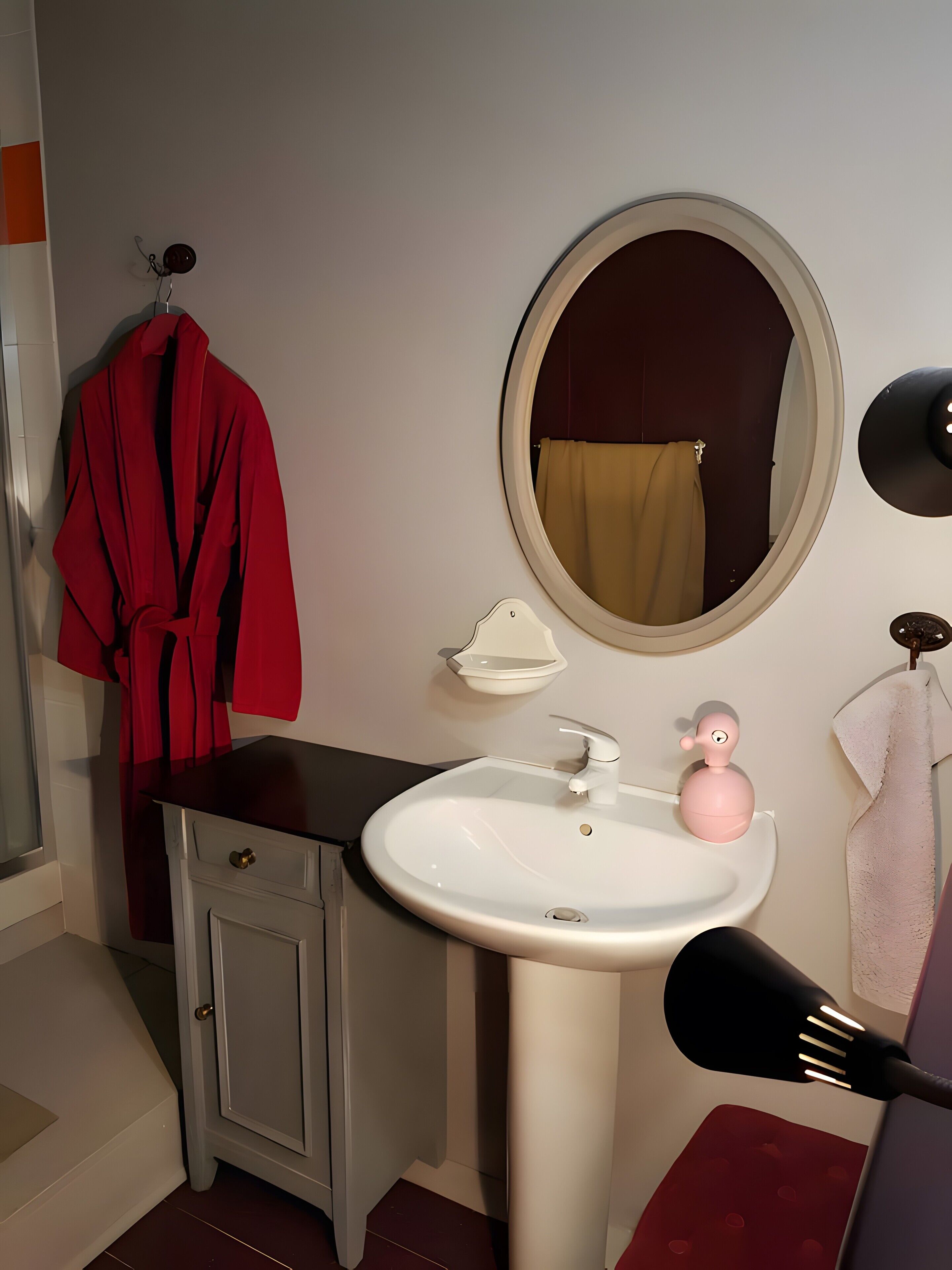 Une salle de bain avec un lavabo blanc, un miroir rond, une robe rouge suspendue et un porte-serviettes.
