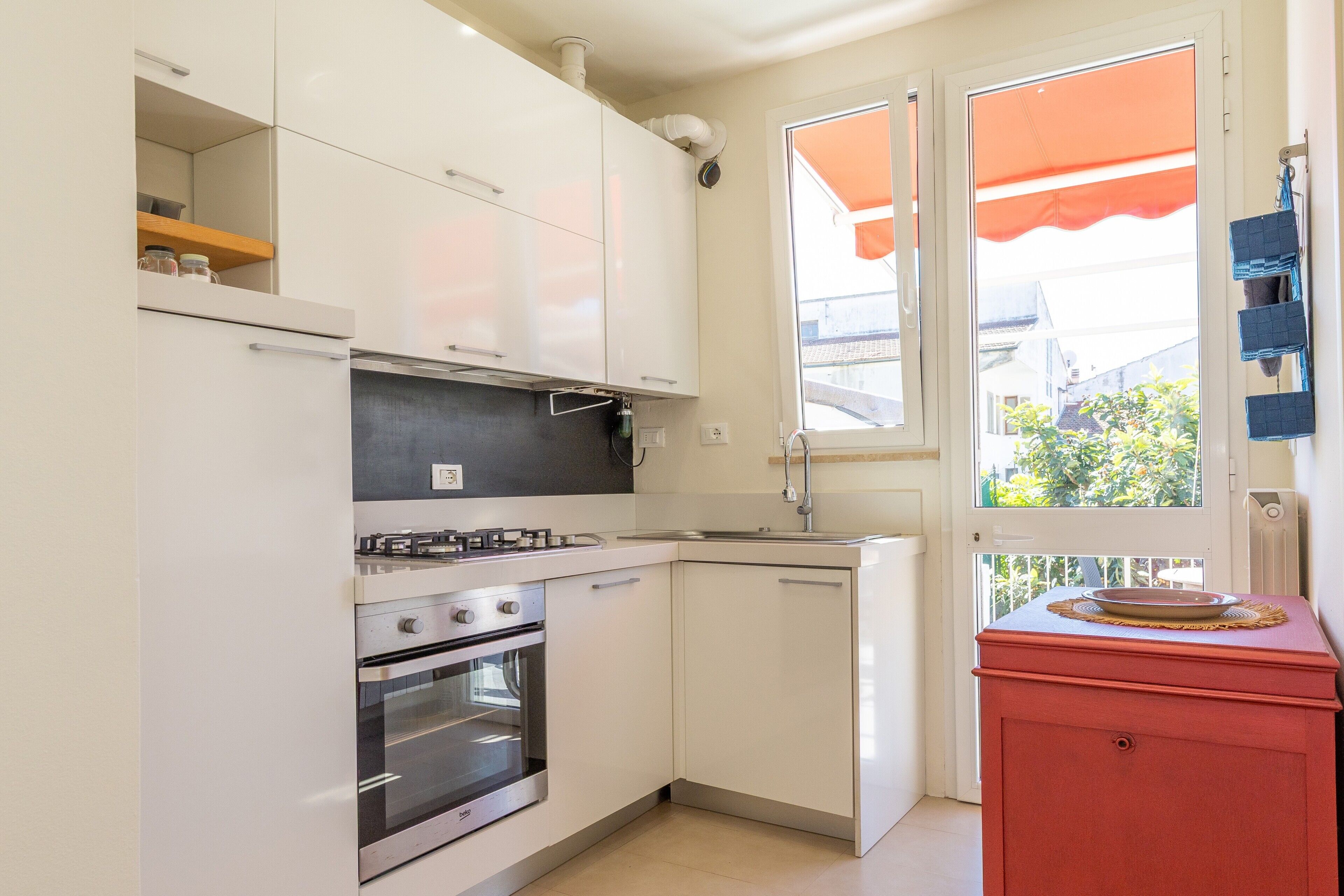 Een kleine keuken met witte kastjes, een rode aanrecht en een raam met een oranje luifel.