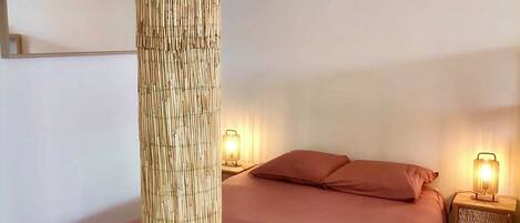 A bedroom with a bed, two bedside tables with lamps, a tall woven column, and a mirror on the wall.