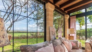 Un espacio al aire libre techado con un sofá y cojines, una lámpara y un cuadro, con vistas a un jardín cercado.