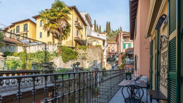 A narrow street with buildings on either side, a metal fence, and a table with chairs on a balcony. - Appartamento The Rich Man (Bellagio)