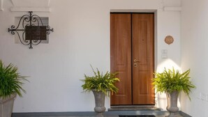 A double wooden door with a metal handle, two potted plants on either side, a wall-mounted sign, and a doormat on the floor. - Appartamento The Rich Man (Bellagio)