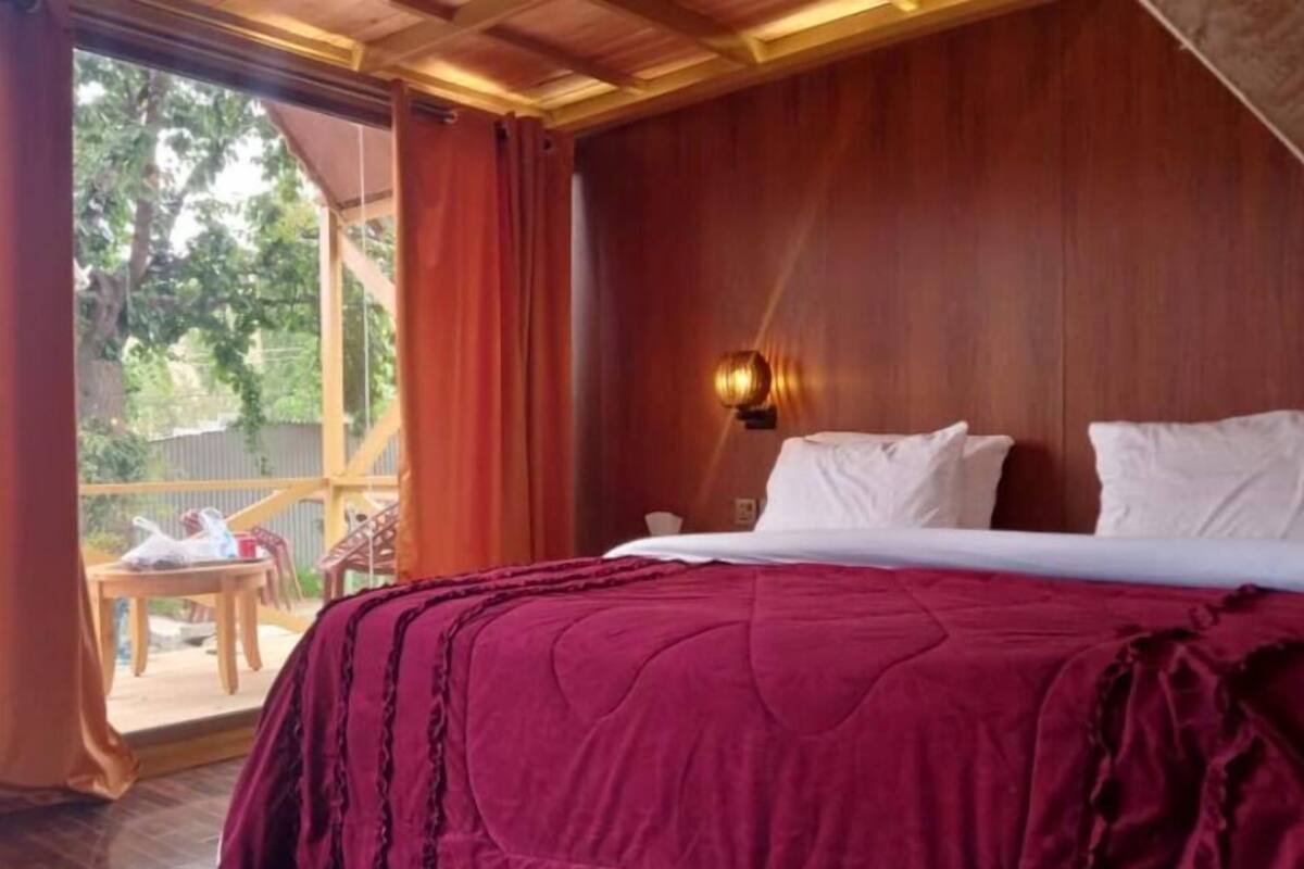 A bedroom with a wooden ceiling, a bed with a red blanket, white pillows, and a nightstand with a lamp.