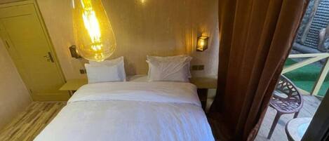 Habitación de hotel con cama, lámpara colgante, mesita de noche, lámpara de pared y vistas al exterior.