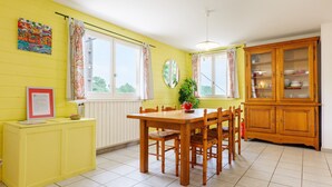 Ein Essbereich mit einem Holztisch und Stühlen, gelben Wänden, einem Fenster mit Vorhängen, einer Speisekammer mit Geschirr und einer Pflanze.