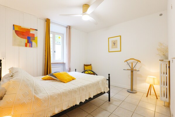 A bedroom with a bed, a bedside table, a hanging light, a window with curtains, and a wall with a picture.