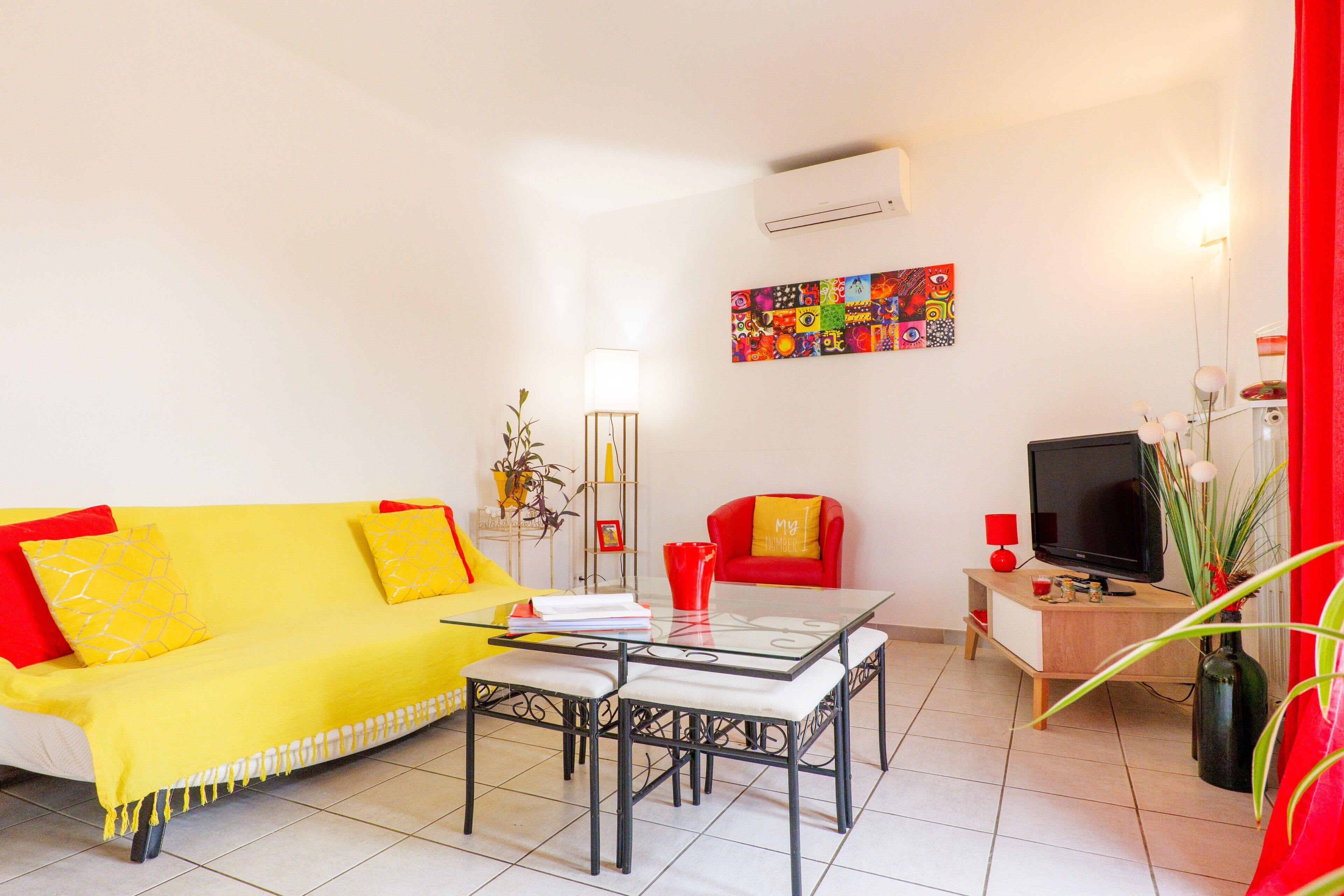 A living room with a yellow sofa, a glass coffee table, a red armchair, a television, and a colorful abstract painting on the wall.