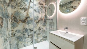 A bathroom with a marble wall, a glass shower enclosure, a white sink, and a round mirror with a light. - Companies, large families, free parking (ROUEN)
