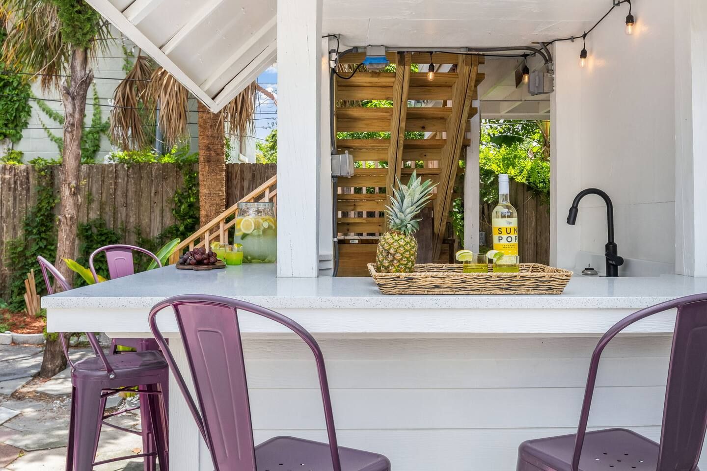 An outdoor bar area with a white countertop, purple chairs, and a wooden shelf with drinks and a pineapple.