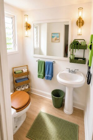 Ein Badezimmer mit Toilette, Waschbecken, einer grünen Fußmatte, einem Korbwäscheschrank und einem grünen Haartrockner an der Wand.