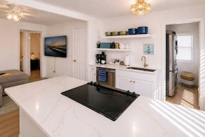 A modern kitchen with a central island, a dining area with a sofa, and a view of a sunset through a window. - Historic Uptown St. Pete 2BR | Fenced Yard + Wet Bar | Walk to Round Lake (Saint Petersburg)