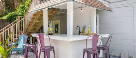 A covered outdoor bar area with a wooden staircase leading to a second floor, two purple bar stools, and a bottle of liquor on the counter.