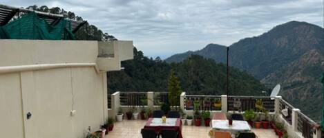 A rooftop terrace with tables and chairs, potted plants, and a view of mountains.