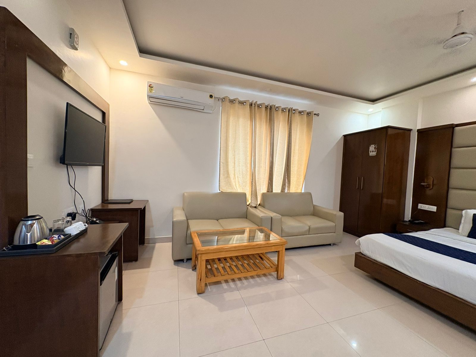 A hotel room with a bed, two sofas, a wooden coffee table, a TV, a desk with a kettle, and a wardrobe.