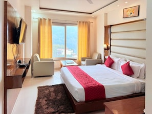 A hotel room with a bed, two chairs, a desk, a TV, and a window with curtains. - Hotel Bliss Ganga  (Rishikesh)