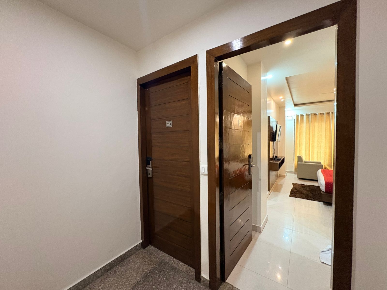 Un pasillo con una puerta de madera marcada '314' que da a una habitación con una cama, un televisor y una ventana con cortinas.