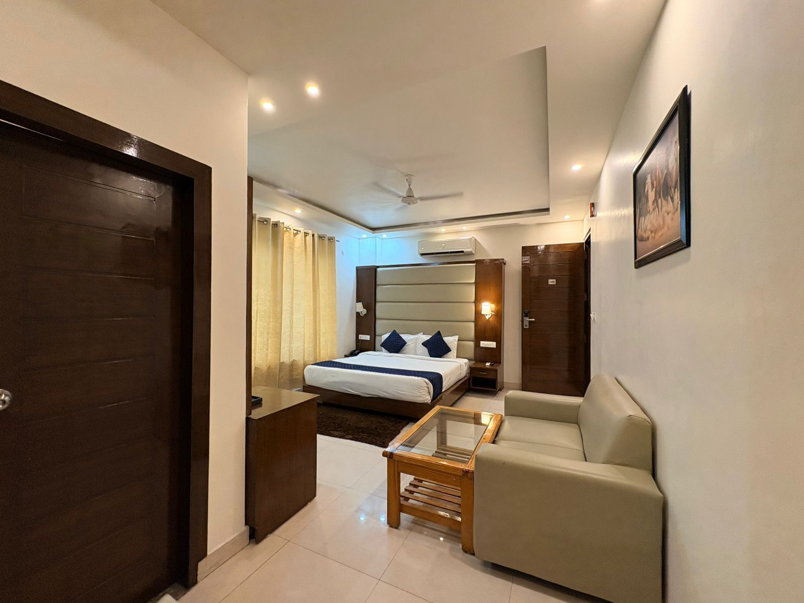A hotel room with a bed, a sofa, a small table, a ceiling fan, and a wall-mounted air conditioner.