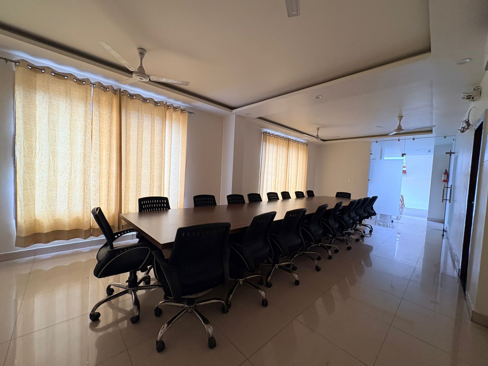 Un salón de conferencias con una larga mesa, múltiples sillas, ventiladores de techo y ventanas grandes con cortinas.