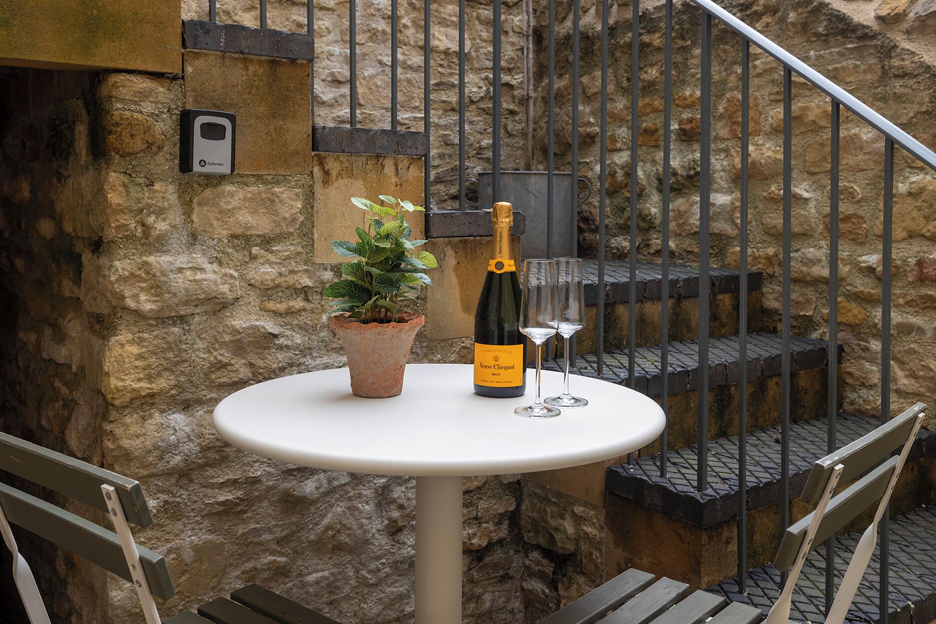 Una mesa con una botella de Veuve Clicquot Brut champagne y dos copas, una planta en maceta y una pared de piedra con una escalera de metal.