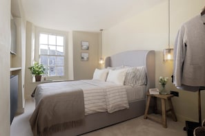A bedroom with a bed, a window, a bedside table, and a hanging garment. - Church Alley House (Cheltenham)